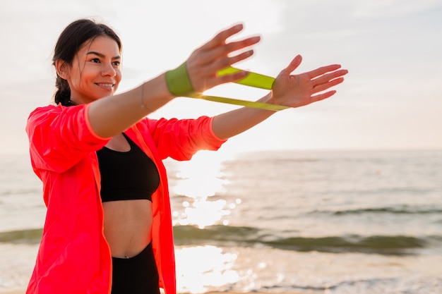 Free photo young attractive woman doing sport exercises in morning sunrise on sea beach, healthy lifestyle, listening to music on earphones, wearing pink windbreaker jacket, making stretching in rubber band