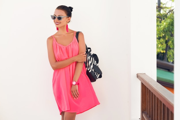 Free Photo young attractive woman in pink dress against wall, holding backpack, wearing hipster sunglasses, fashion accessories, modern street style, summer trend, traveling on vacation, smiling, happy emotion