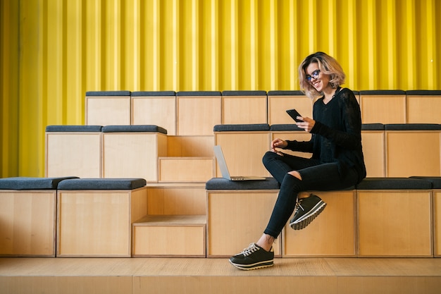 Young attractive woman sitting in lecture hall, working on laptop, wearing glasses, modern auditorium, student education online, freelancer, smiling, using smartphone, digital devices