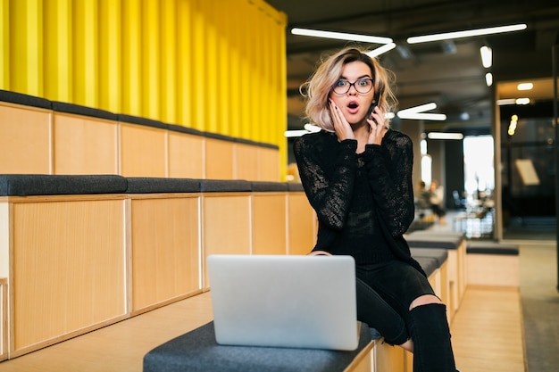 Young attractive woman sitting in lecture hall working on laptop wearing glasses, modern auditorium, student education online, shocked face expression