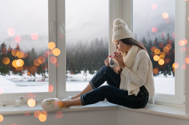 Free photo young attractive woman in stylish white knitted sweater, scarf and hat sitting at home on windowsill at christmas holding cup drinking hot tea, winter forest background view, lights bokeh