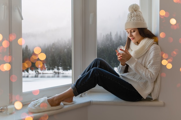 Free photo young attractive woman in stylish white knitted sweater, scarf and hat sitting at home on windowsill at christmas holding glass snow ball present decoration, winter forest view, lights bokeh