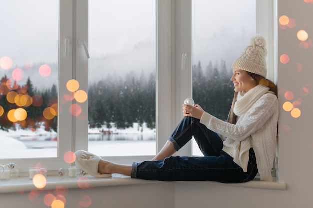 Young attractive woman in stylish white knitted sweater, scarf and hat sitting at home on windowsill at Christmas holding glass snow ball present decoration, winter forest view, lights bokeh