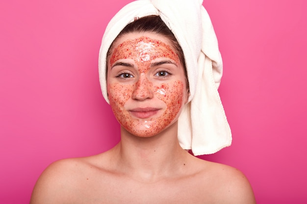 Young attractive woman with white towel on her head, has naked body, smilling isolated over pink wall in studio, looks directly at camera, having red scrub on her face.