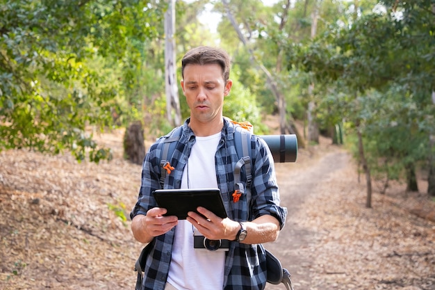 Free photo young backpacker hiking, holding tablet and looking at map. caucasian attractive traveler walking on road in woods. backpacking tourism, adventure and summer vacation concept