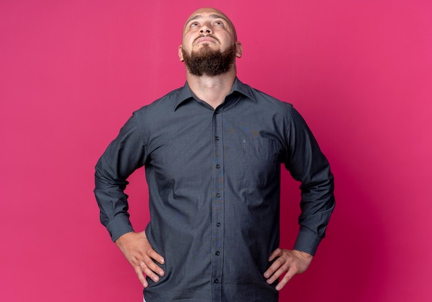 Free Photo young bald call center man putting hands on waist and looking up isolated on crimson background with copy space