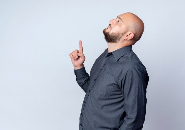 Young bald call center man standing in profile view looking and pointing up isolated on white background with copy space