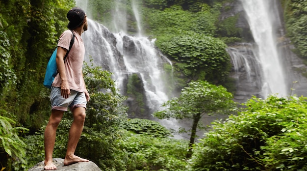 Free Photo young barefooted tourist in baseball cap standing on big stone and looking back at waterfall behind him in beautiful exotic nature. bearded traveler enjoying wildlife while hiking in rainforest