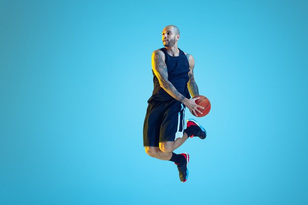 Young basketball player of team wearing sportwear training, practicing in action, motion on blue wall in neon light