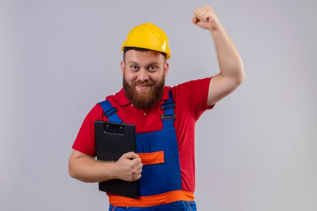 Free photo young bearded builder man in construction uniform and safety helmet holding clipboard raising fist positive and happy, winner concept