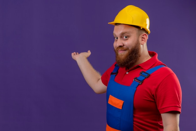 Free photo young bearded builder man in construction uniform and safety helmet smiling confident presenting something behind him with arm of his hand over purple background