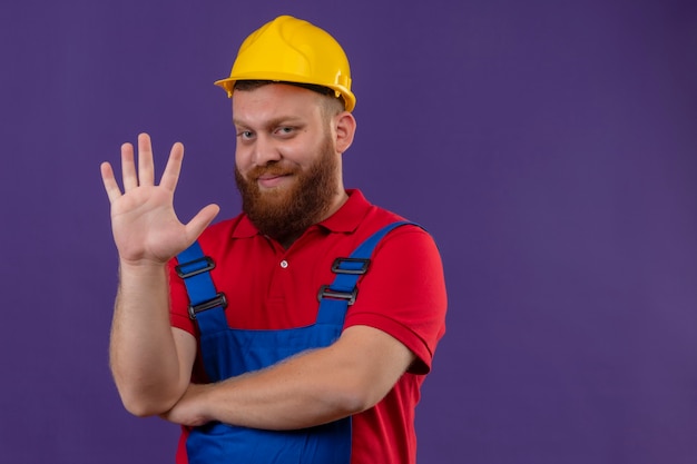 Young bearded builder man in construction uniform and safety helmet smiling showing and pointing up with fingers number five over purple background