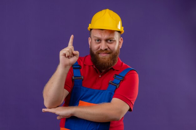 Free Photo young bearded builder man in construction uniform and safety helmet with skeptic expression pointing finger up over purple background