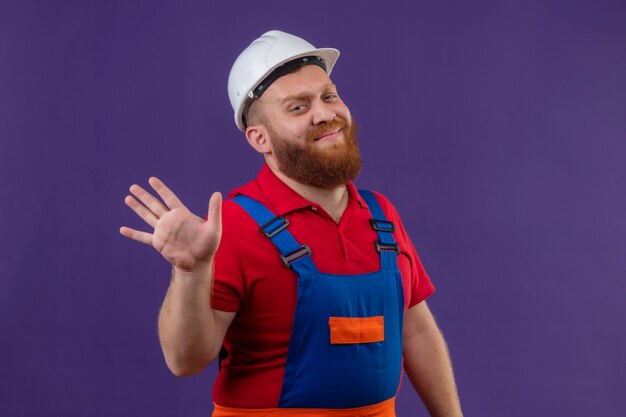 Young bearded builder man in construction uniform and safety helmet with skeptic smile waving with hand over purple background