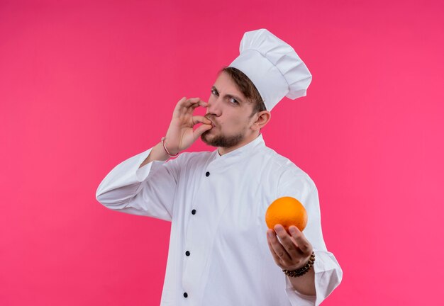 A young bearded chef man in white uniform holding orange and showing tasty ok gesture while looking on a pink wall