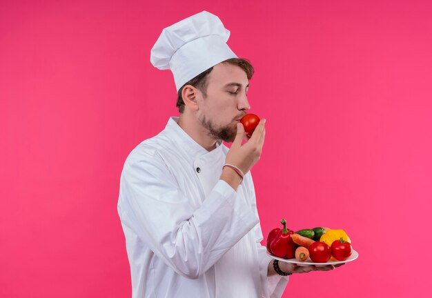 A young bearded chef man in white uniform holding a plate with fresh vegetables while smelling tomato on a pink wall