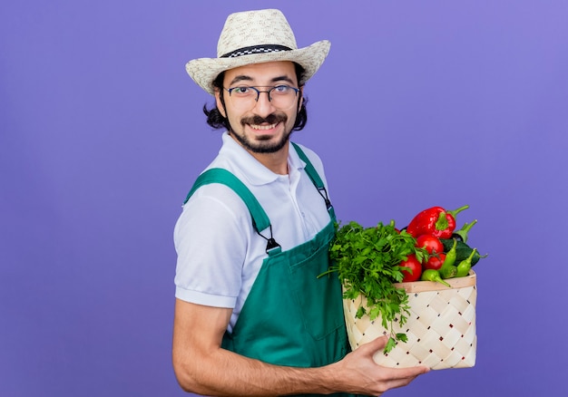 Young bearded gardener man wearing jumpsuit and hat holding crate full of vegetables looking at front smiling cheerfully standing over blue wall