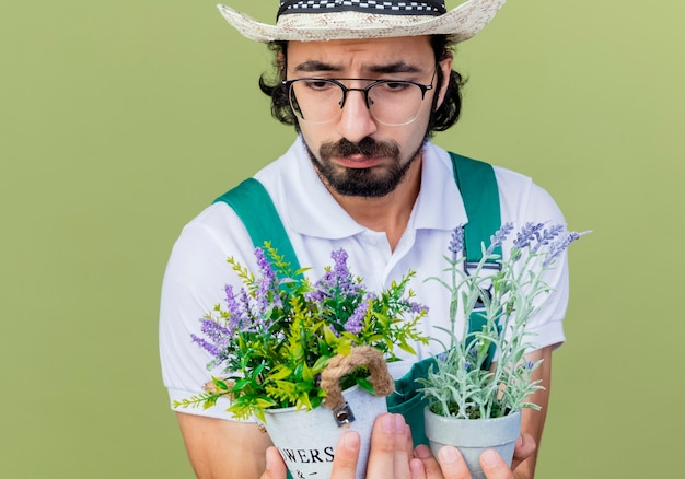 Young bearded gardener man wearing jumpsuit and hat holding potted plants looking at them with sad expression standing over light green wall