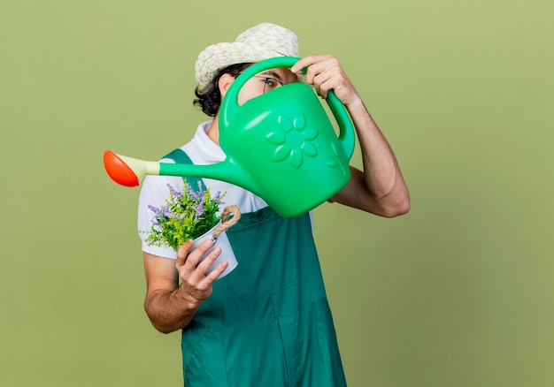 Free Photo young bearded gardener man wearing jumpsuit and hat holding watering can and potted plant hiding face standing over light green wall