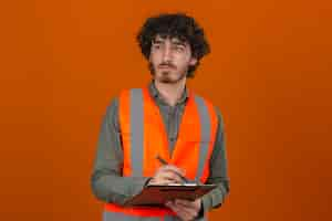 Free photo young bearded handsome engineer wearing construction vest holding clipboard writing something looking aside thoughtful expression on face over isolated orange wall