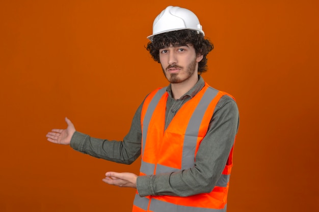 Free photo young bearded handsome engineer wearing security helmet and vest presenting and pointing with palms of hands looking at the camera with serious face over isolated orange wall