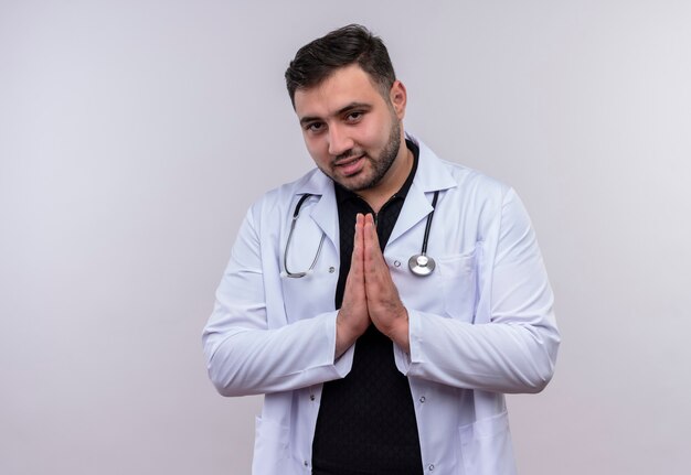 Free photo young bearded male doctor wearing white coat with stethoscope holding hands together in prayer gesture feeling thankful and happy