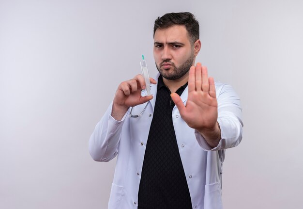 Young bearded male doctor wearing white coat with stethoscope holding syringe  making stop sign with hand with serious frowning face 