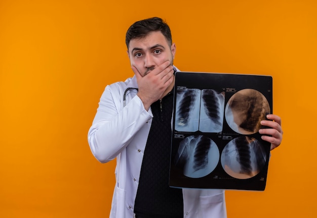 Free photo young bearded male doctor wearing white coat with stethoscope holding x-ray of lungs shocked covering mouth with hand