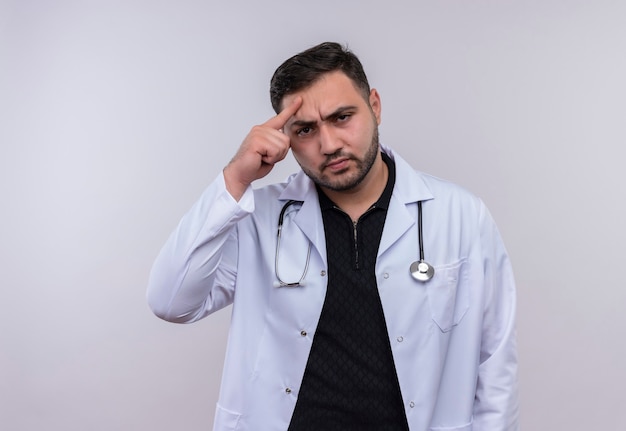 Young bearded male doctor wearing white coat with stethoscope looking at camera with serious face displeased 