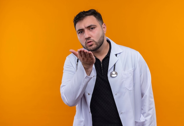 Young bearded male doctor wearing white coat with stethoscope raising arm gesturing with hand as asking question 