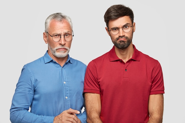 Young bearded male raises eyebrow in bewilderment, dressed in red t-shirt, stands next to his mature father, spend weekend in family circle, isolated over white wall. Relationship concept