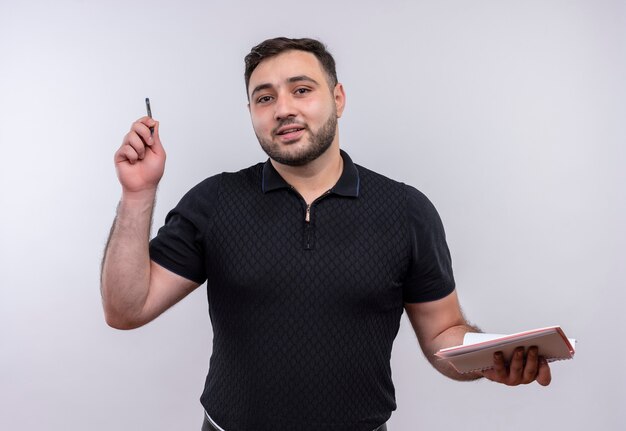 Young bearded man in black shirt holding notebook and pen looking confident 