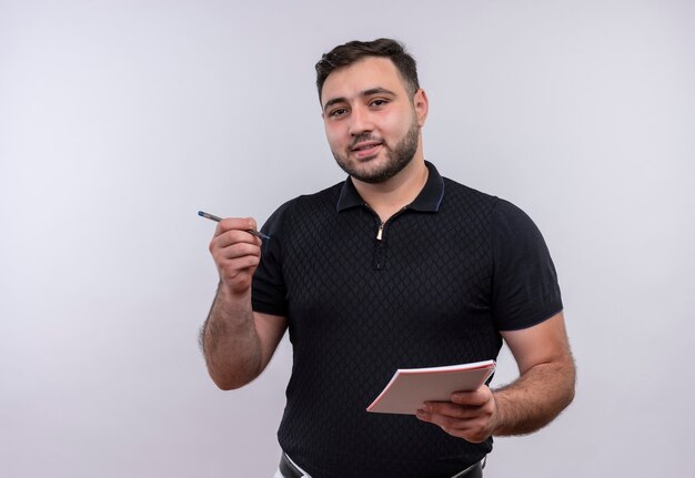 Young bearded man in black shirt holding notebook and pencil smiling 