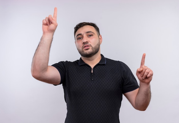 Young bearded man in black shirt pointing index fingers up with confident serious expression 