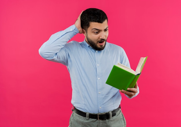 Young bearded man in blue shirt holding book looking at it being surprised and confused 