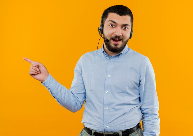 Young bearded man in blue shirt with headphones with microphone pointign with index finger to the side smiling 