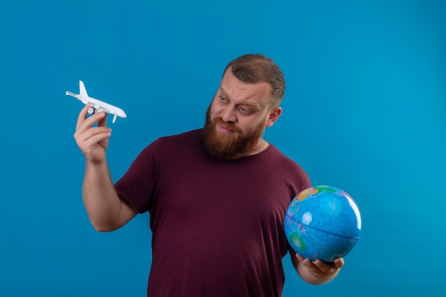 Free photo young bearded man in brown t-shirt holding globe and toy airplane looking at toy with skeptic expression on face