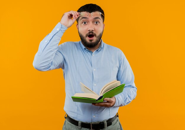 Young bearded man in glasses and blue shirt holding book being amazed putting his glasses off 