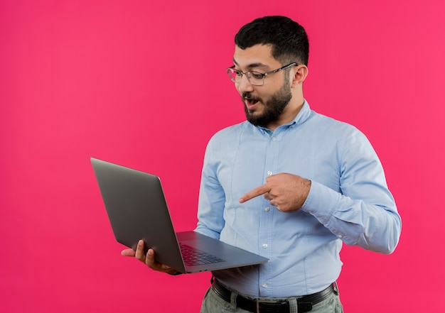 Young bearded man in glasses and blue shirt holding laptop pointing with finger at it being surprised 