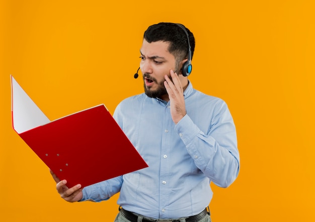 Free photo young bearded man in glasses and blue shirt with headphones with microphone holding big folder looking at it surprised and amazed