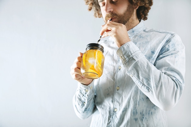 Free Photo young bearded man in light jeans shirt drinks fresh home made orange citrus sparkling lemonade through striped drinking straw from rustic transparent jar in hands