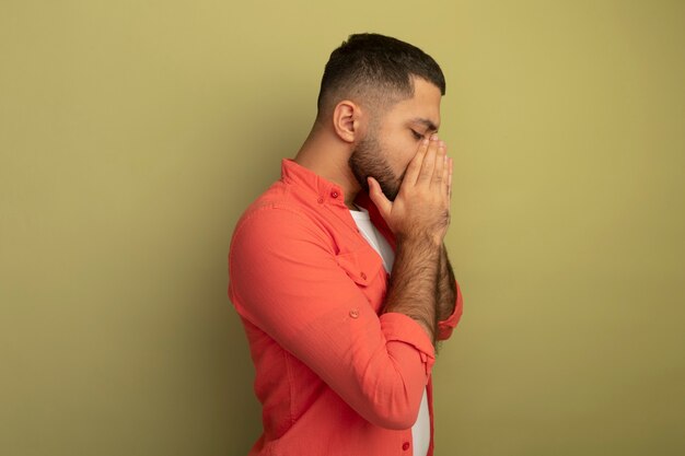 Young bearded man in orange shirt standing sideways with hands on face depressed and sad over light wall