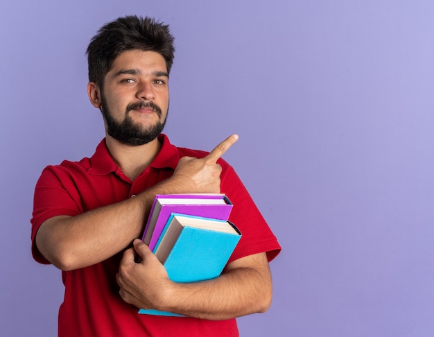 Young bearded student guy in red polo shirt holding books looking confident smiling pointing with index finger to the side standing