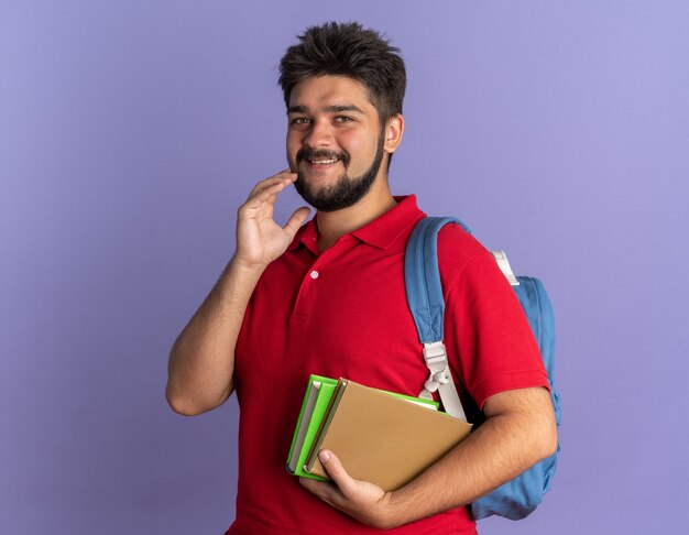 Young bearded student guy in red polo shirt with backpack holding books  with shy smile on face standing over blue wall