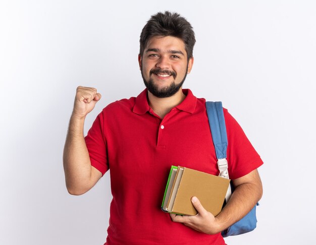 Young bearded student guy in red polo shirt with backpack holding notebooks lookign  happy and excited clenching fist standing over white wall