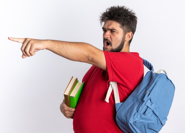 Young bearded student guy in red polo shirt with backpack holding notebooks looking aside with angry face shouting pointing with index finger at something standing over white wall