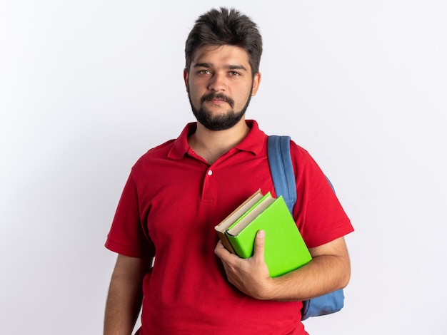 Young bearded student guy in red polo shirt with backpack holding notebooks looking at camera with serious confident expression standing over white background