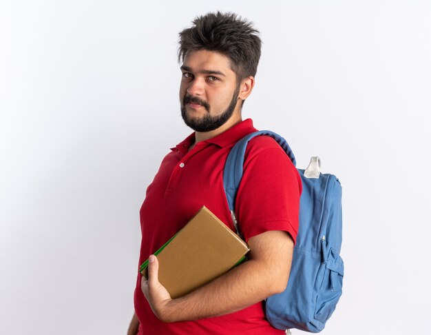 Young bearded student guy in red polo shirt with backpack holding notebooks looking smiling happy and positive standing