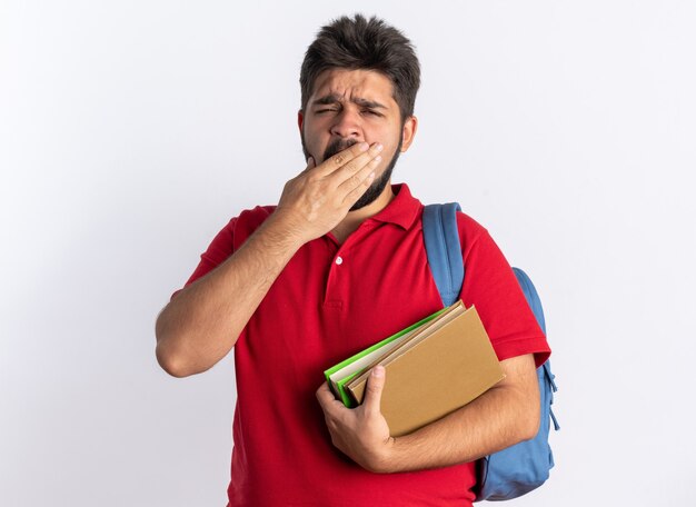 Young bearded student guy in red polo shirt with backpack holding notebooks looking tired and bored yawning standing over white background