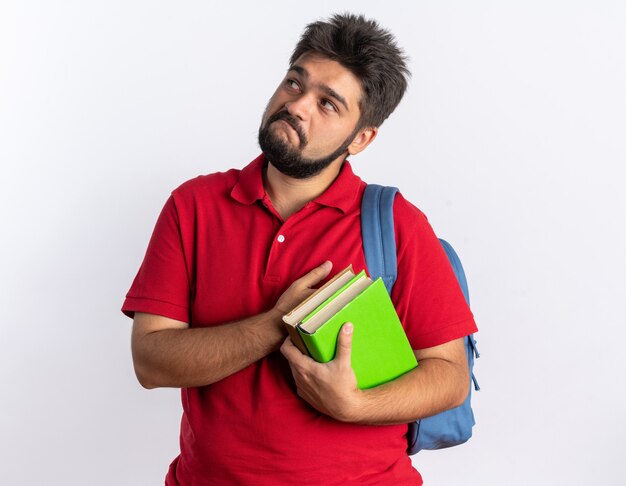 Young bearded student guy in red polo shirt with backpack holding notebooks looking up with skeptic expression on face standing over white background